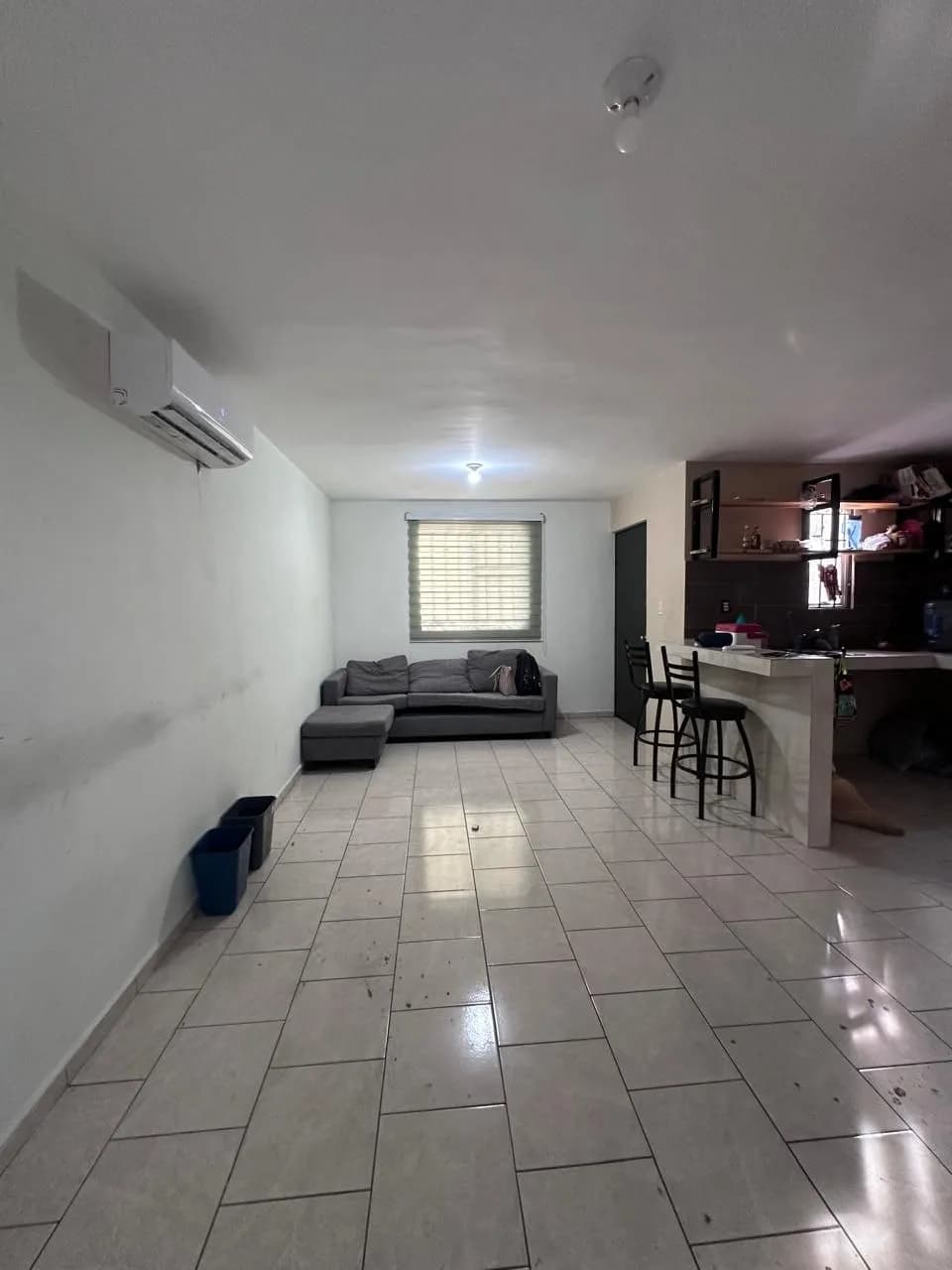 Sala con sofá gris, ventana con persiana, aire acondicionado y barra de cocina al fondo