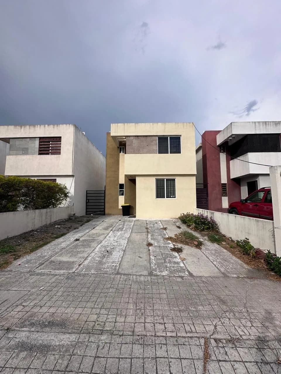 Casa de dos pisos beige con cochera descubierta entre otras viviendas bajo cielo nublado